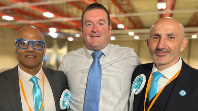 Karan Maheshwari, Ryan Coogan and Julian Tobias are all standing side by side. Maheshwari is wearing blue glasses, a white shirt, blue tie and grey blazer. He is wearing a Reform UK rosette. Coogan has short brown hair and is wearing a blue and white stripped T-shirt and blue tie. Tobias is wearing a white shirt, blue tie and black suit jacket. He is also wearing a Reform UK rosette and sticker.