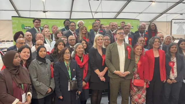 A group of councillors wearing red rosettes stand in several rows posing for a photo