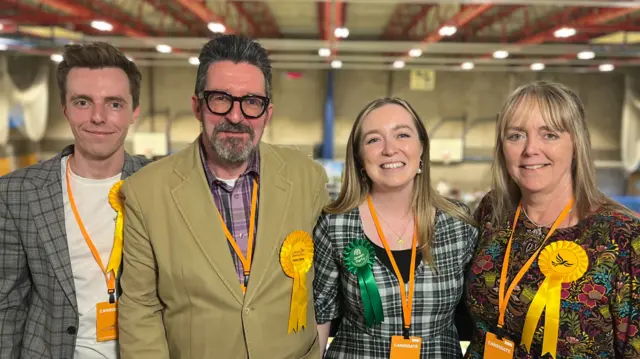 A family are four are standing next to each other and are smiling. From left to right is Nathan, Alan, Georgie, and Jacqueline. Nathan is wearing a white T-shirt and chequered blazer. He is also wearing a Liberal Democrat rosette. Alan is wearing a purple chequered shirt and a green coat, he is also wearing black framed glasses and a Liberal Democrat rosette. Georgie has long blonde hair and is wearing a black and white dress, a Green Party rosette and a lanyard. Jacqueline is wearing a floral dress and a Liberal Democrats rosette. She has long blonde hair and a fringe.