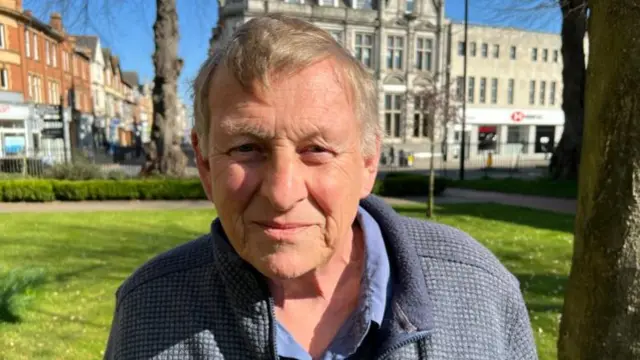 A man in a blue fleece and shirt with fair hair and eyebrows looks into camera with a grassy area and high street behind him.