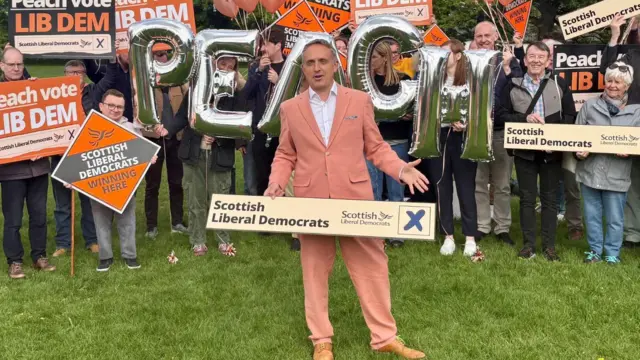 Alex Cole-Hamilton holding a Scottish Liberal Democrats banner wearing a peach suit and standing in front of supporters holding placards