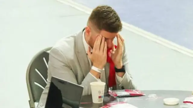 A man is holding his head in is hands, looking down at a table