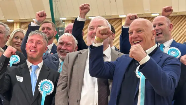 A group of Reform candidates have their fists aloft in celebration, they are smiling as if they are mid-cheer and wearing turquoise rosettes/