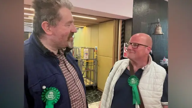 Two men wearing green rosettes are smiling at each other. The taller man, on the left, has short brown hair and is wearing a checked shirt and blue jacket. The man on the right is bald and is wearing glasses. He is wearing a white body warmer.