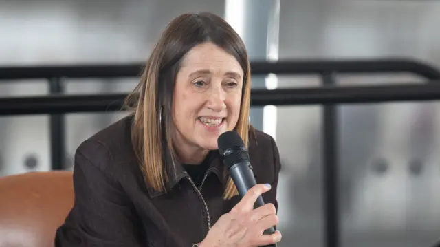 A woman is sitting down and leaning forward, holding a microphone towards her face