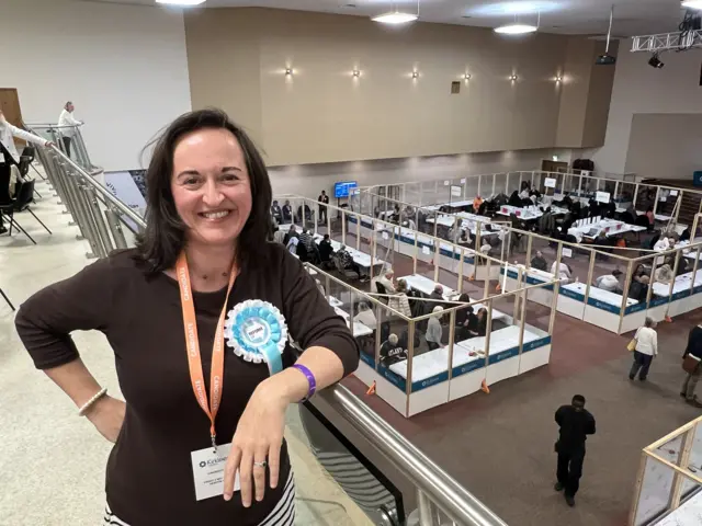 Councillor Sarah Wood standing on a balcony overlooking people counting votes