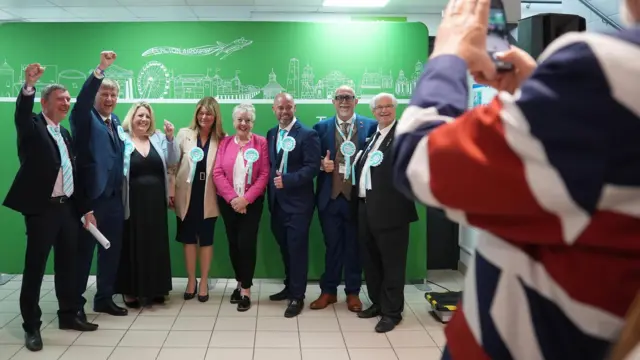 A group of people dressed smartly and wearing rosettes cheer, smile and do a thumbs up while being photographed by someone draped in a flag