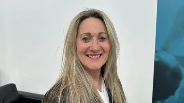 A woman with long blonde hair smiles at the camera. She wears a black suit jacket with a white top underneath.
