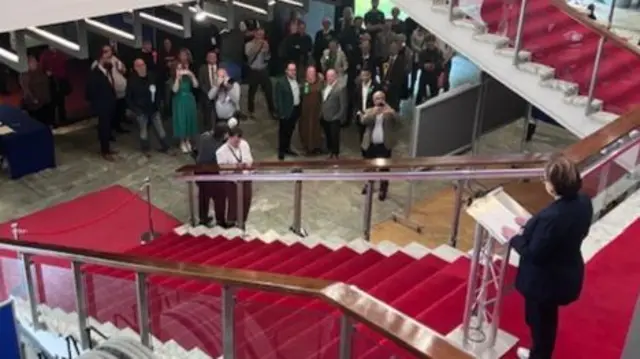 A woman stands on the at the top of a red-carpeted stairway, reading from a podium. There is a crowd of people gathered below, looking up at her
