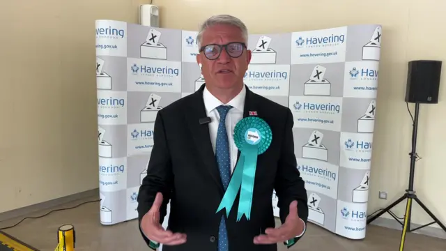 A man in a dark suit with a Reform UK rosette is being interviewed for TV.