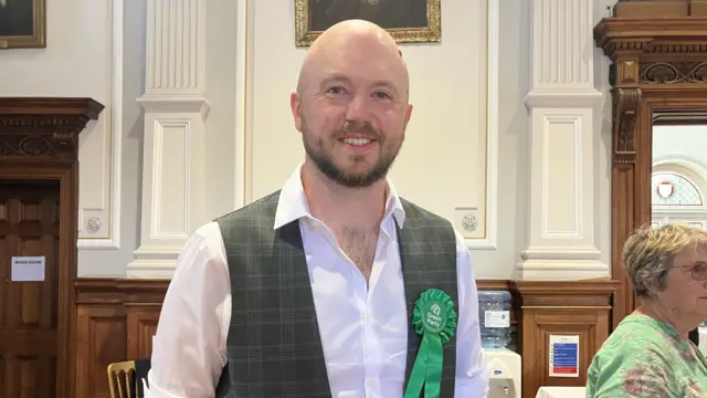 A man in a white shirt and waistcoat wearing a Green Party rosette. He is smiling and has a bald head and a brown beard