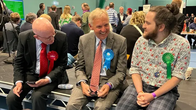 Three men wearing rosettes share a joke and are all pictured laughing or smiling