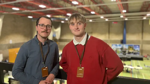 Phil Shepka and Finn Cullum are standing next to each other on a balcony. Shepka has short brown hair and is wearing a grey shirt and a brown lanyard. Cullum is on the right and has short blonde hair and is wearing a white shirt and red jumper.