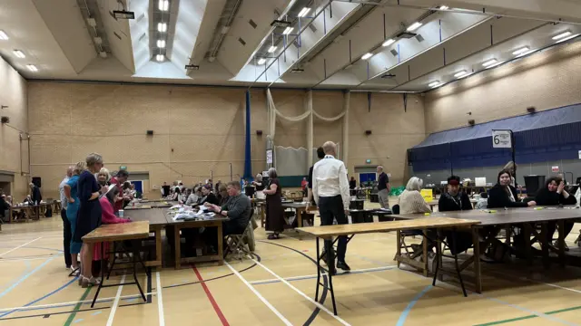 Long tables laid out in a sports hall. People are sitting around them, some sorting through papers