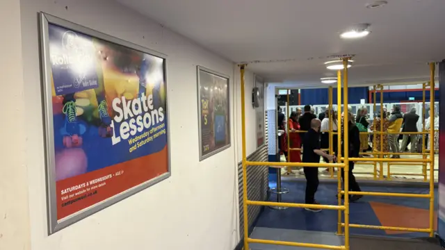 The entrance to Roller City, with a board advertising Skate Lessons to the left and yellow railings to the right, with people gathered for a count beyond.
