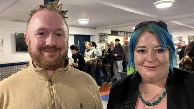 Larry Crofton with brown hair tied at the back, and a short beard, wearing a brown fleece, and Lucy Musk, with medium-length blue hair wearing a black sweater over a pink top with a blue necklace. There is a queue of people heading for lunch behind them.