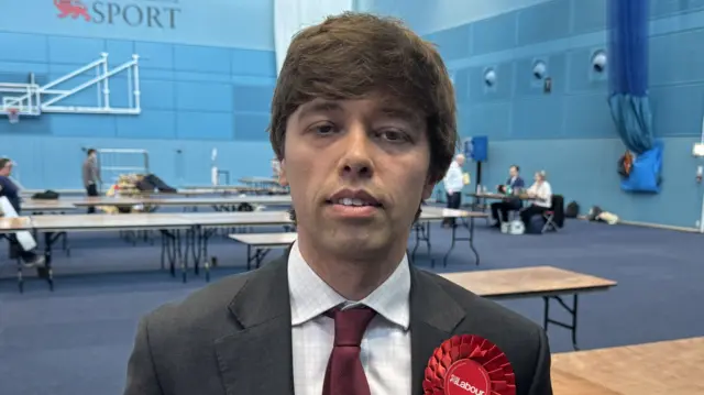 Cameron Holloway is in the middle standing in a room which has several tables. He has short brown hair and is wearing a white chequered shirt, red tie and a grey suit jacket which has a rosette on it.