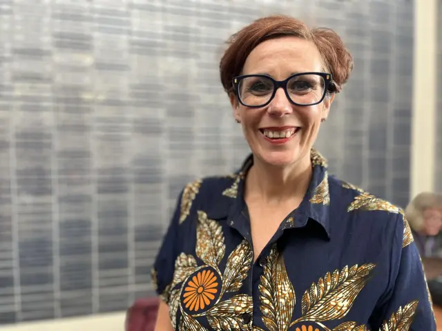 Sam Mowbray smiles at the camera. She wears glasses and a colourful patterned dress.