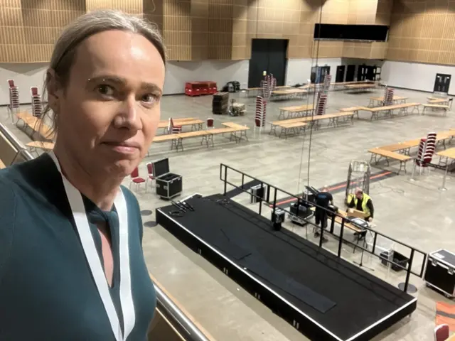 A woman takes a selfie on the balcony as the staging of the count is packed down behind her. she is wearing a white lanyard and green top.