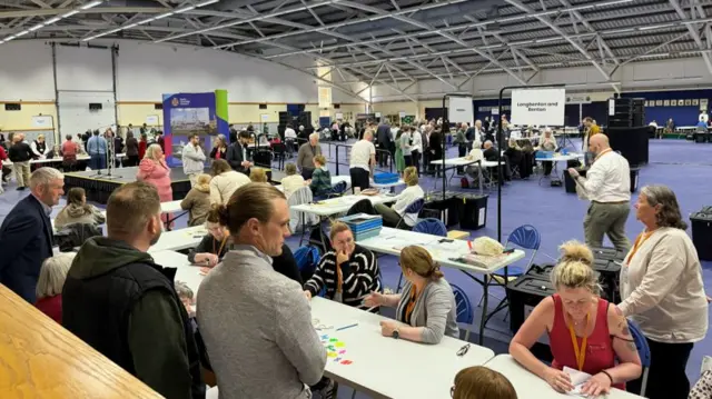 The North Tyneside count in the large sports hall, which has a purple carpet and yellow and white walls. There are tables dotted around the room, with people sat at them riffling through ballot papers. There are also people standing on the other side observing.