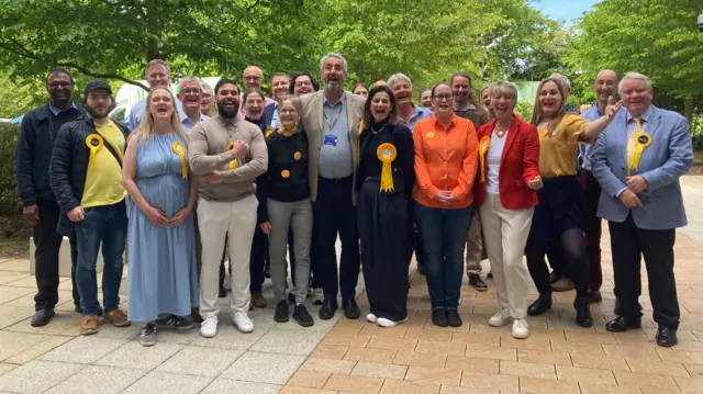 Several people are standing in a group cheering. They are all wearing Liberal Democrat rosettes and are standing on a pathway.