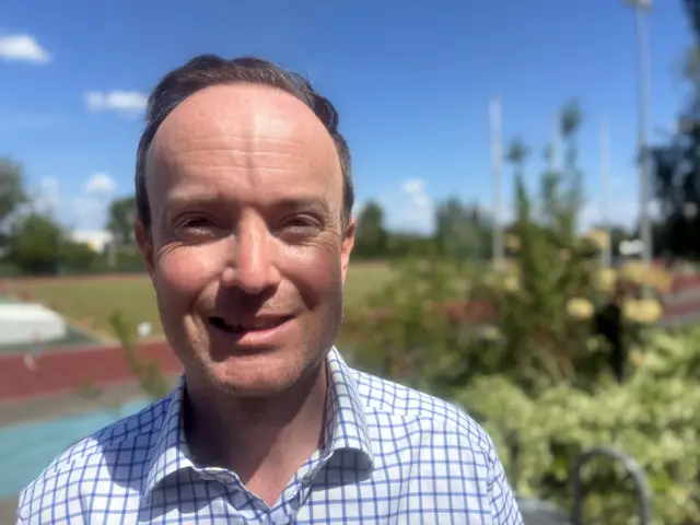 A man with short brown hair smiles at the camera while standing outside in the sun. He wears a white and blue chequered shirt.