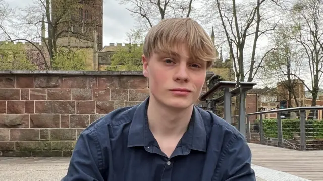 Ben has blond hair and is wearing a dark blue shirt. He is sitting on some steps in front of a low sandstone wall, which itself is in front of a church.