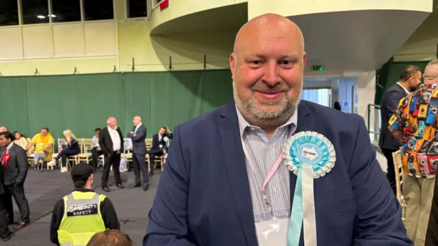 A man wearing a suit and a large rosette smiles at the camera