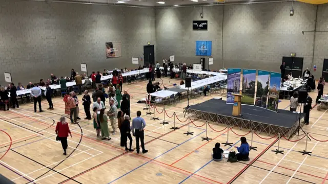 A sports hall with long tables laid out, people standing around and people seated at the tables looking down at papers