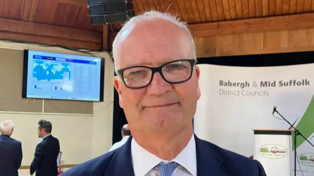 A man with thin grey hair, glasses, wearing a navy suit and white shirt with a blue shirt smiles at the camera.