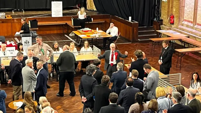 An election count where a man in the centre of the image celebrates after winning.