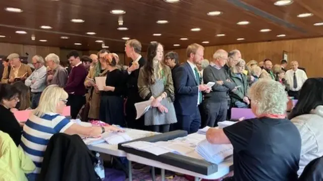A corner table at the election count. There are people sat at a white corner table working through piles of ballot papers. The crowd of people standing in front of them, on the other side of the table, is dense.