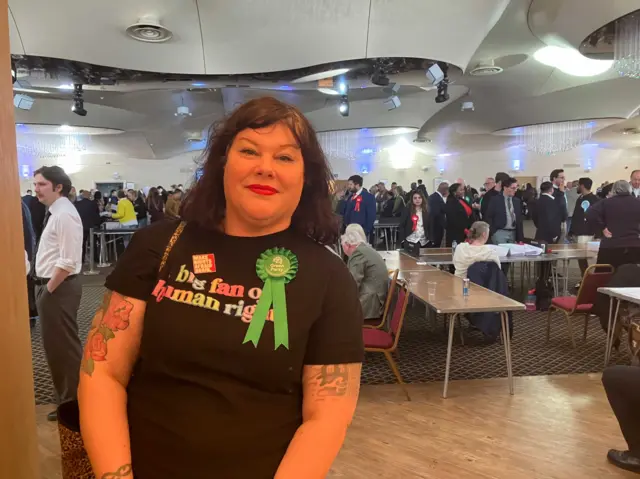 A lady with a Green rosette standing in a large room while vote counting goes on around her