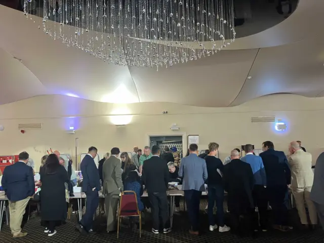 A group of men with their back to the camera stand around tables where votes are being counted.