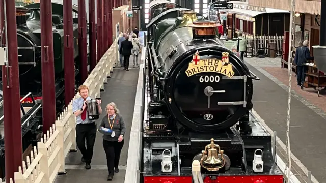 A steam locomotive in a covered museum, with mock platforms down either side. Two people are walking towards the photographer.