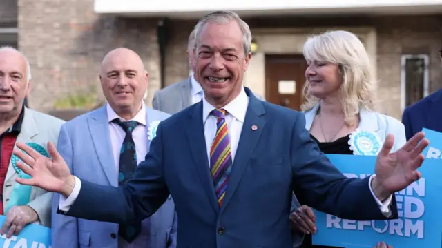 Farage stands holds his arms in a wide open gesture with a big grin on his face as he stands in front of Reform councillors