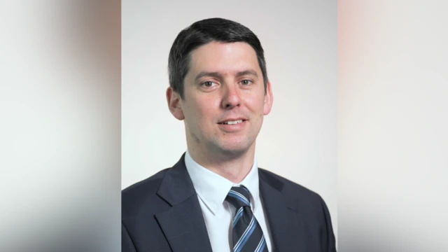 A portrait photo of Chris Chambers. He smiles at the camera. He has short black hair and wears a navy suit with a white shirt and a black, blue and white tie.