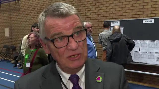 Martin Gannon is speaking to an interviewer behind the camera. He has short grey hair and black glasses. He is wearing a grey suit and a small green badge with a magenta heart on it.