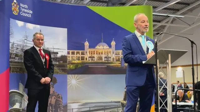 A person in a blue suit and a turquoise rosette for Reform UK stands at the podium.