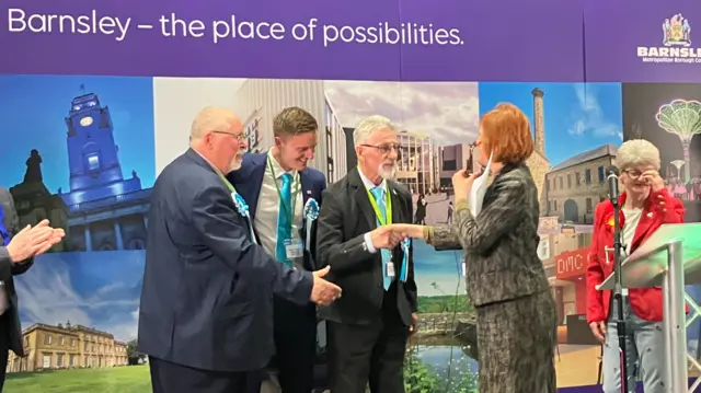 Men in turquouise ties shake hands with the council returning officer, a lady with a ginger bob haircut.