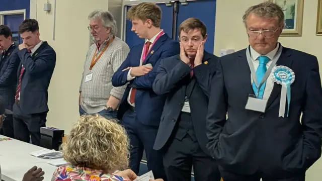 A row of men are looking down at the table where people are counting ballot papers. Second on the right, a young man has both of his hands on his face and a drawn look on hisface.