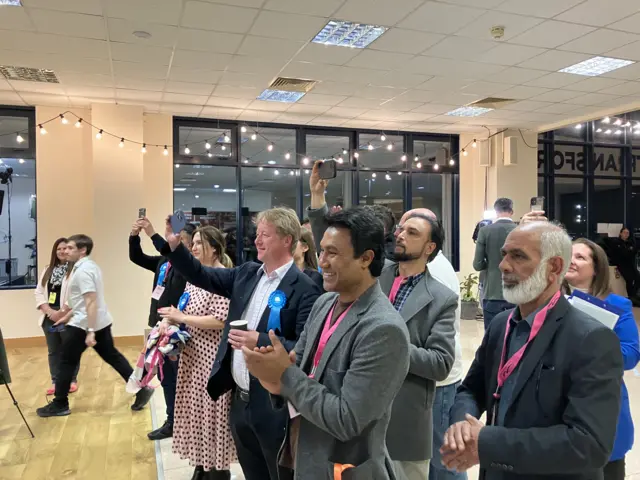 Several people are standing facing the left. Three men in the foreground are wearing blazers and have pink lanyards on. In the background there are more people who are wearing blue rosettes.
