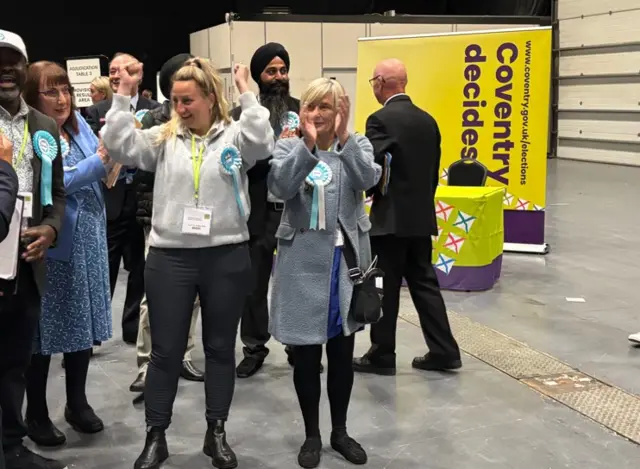 Two women celebrate, the one on the left has long blonde hair and is wearing a grey jumper. She has her arms in the air. The other is in a long blue coat and has short blonde hair. They are in a hall surrounded by others.