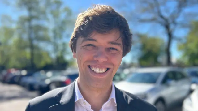 Cameron Holloway is standing in a car park. He has short brown hair and is wearing a white shirt and black blazer. He is smiling.