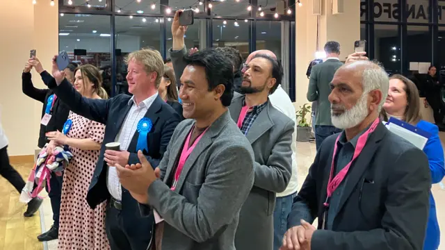 Several people are standing in a group looking to the left. Three men in the foreground are all wearing blazers and pink lanyards. Several people in the background are wearing blue rosettes.