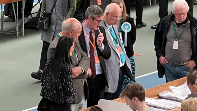 Michael Mordey is among a group of people standing over the table where people are counting ballot papers. He is wearing a dark suit and has short, greying hair. He is holding his pen in his mouth in a pensive way.