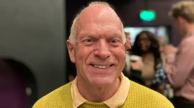 Stephen Giles-Medhurst wearing a yellow jumper is smiling at the camera.