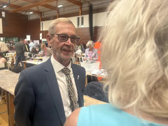 A man in a navy suit jacket and colourful tie with yellow hair and tinted glasses standing in a sports hall with a count going on behind him.