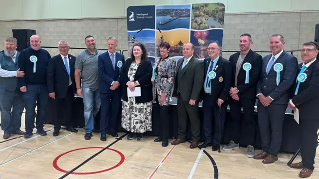 The winning candidates from Reform UK, some wearing blue rosettes, stand in a line in front of a stage at a leisure centre where the election count has taken place. They are mostly men, with two women in the middle of the line-up. They are all smiling.