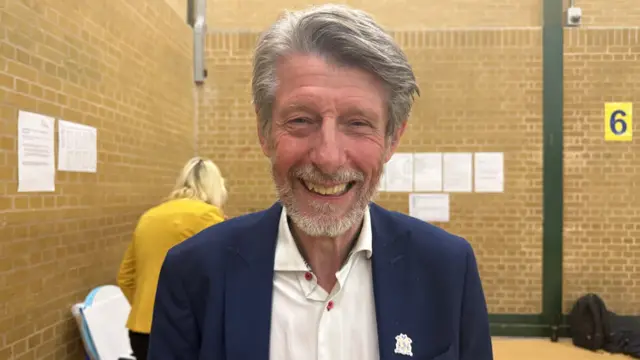 Phil Munday, a man with grey hair and wearing a suit, while stood in a hall and smiling at the camera.